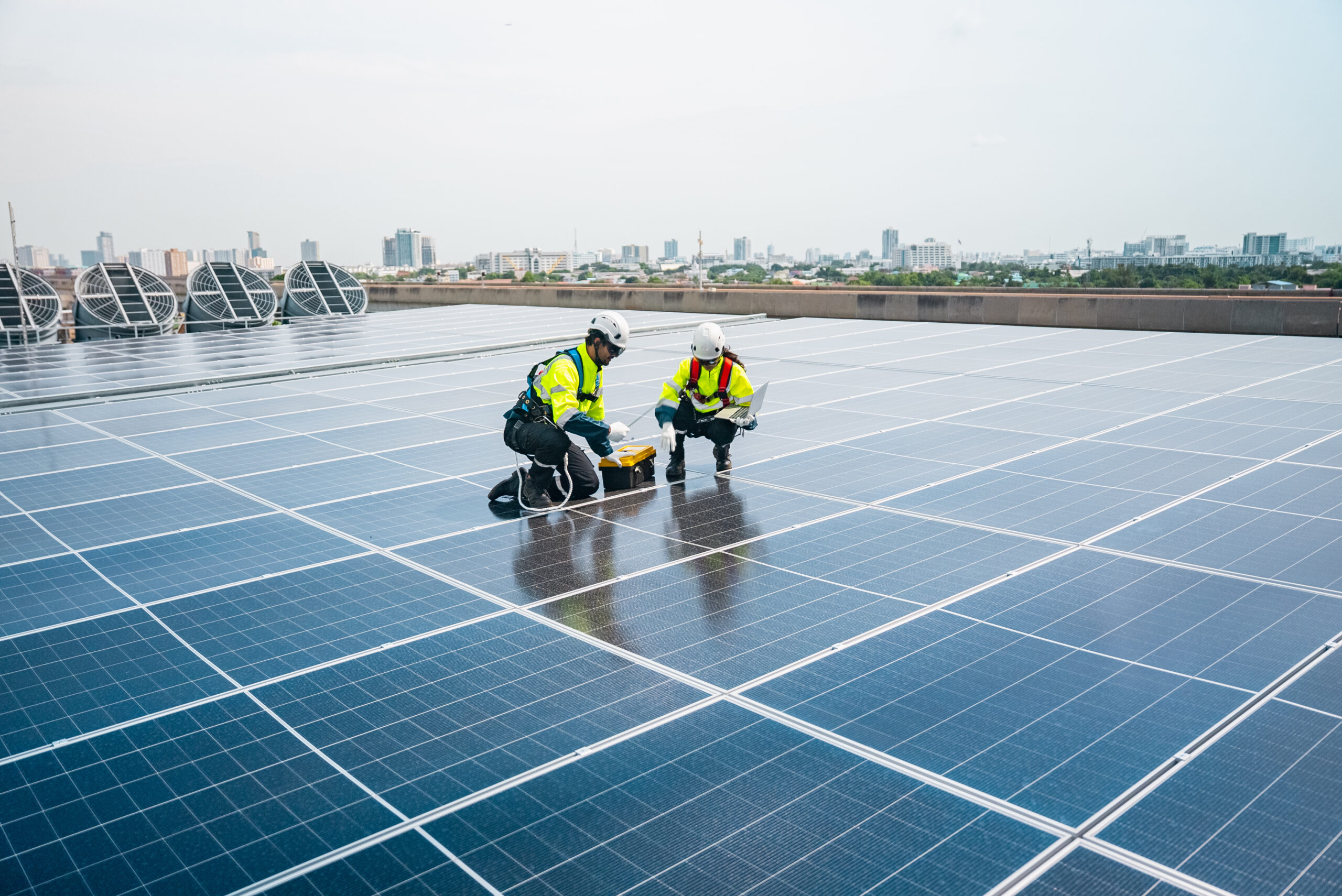Together, a man and female technician wearing luminous vests and safety helmets effectively install solar panels on a sunny day by utilising a laptop and volt meter.